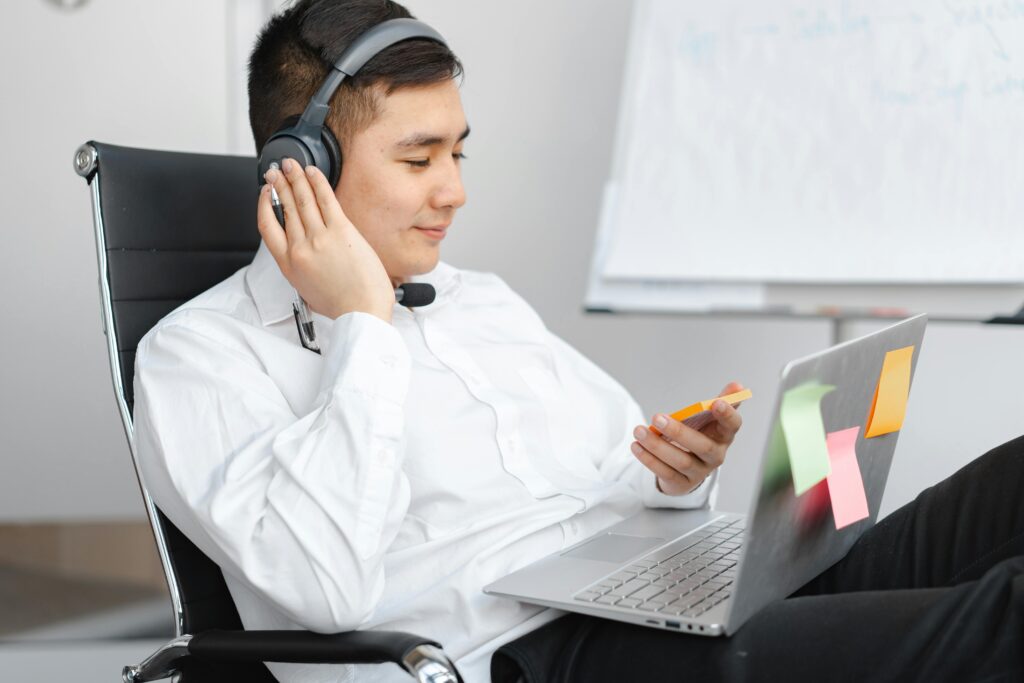Man in White Long Sleeves Sitting on Office Chair while Wearing a Headphone in Front of His Laptop